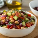 Close-up of protein-packed dense bean salad recipe with black beans, chickpeas, kidney beans, fresh vegetables, herbs, and feta cheese in white bowl