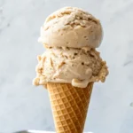 Two scoops of creamy Jamaican Grape Nut Ice Cream served in a waffle cone, showing visible cereal bits and a rich vanilla color.