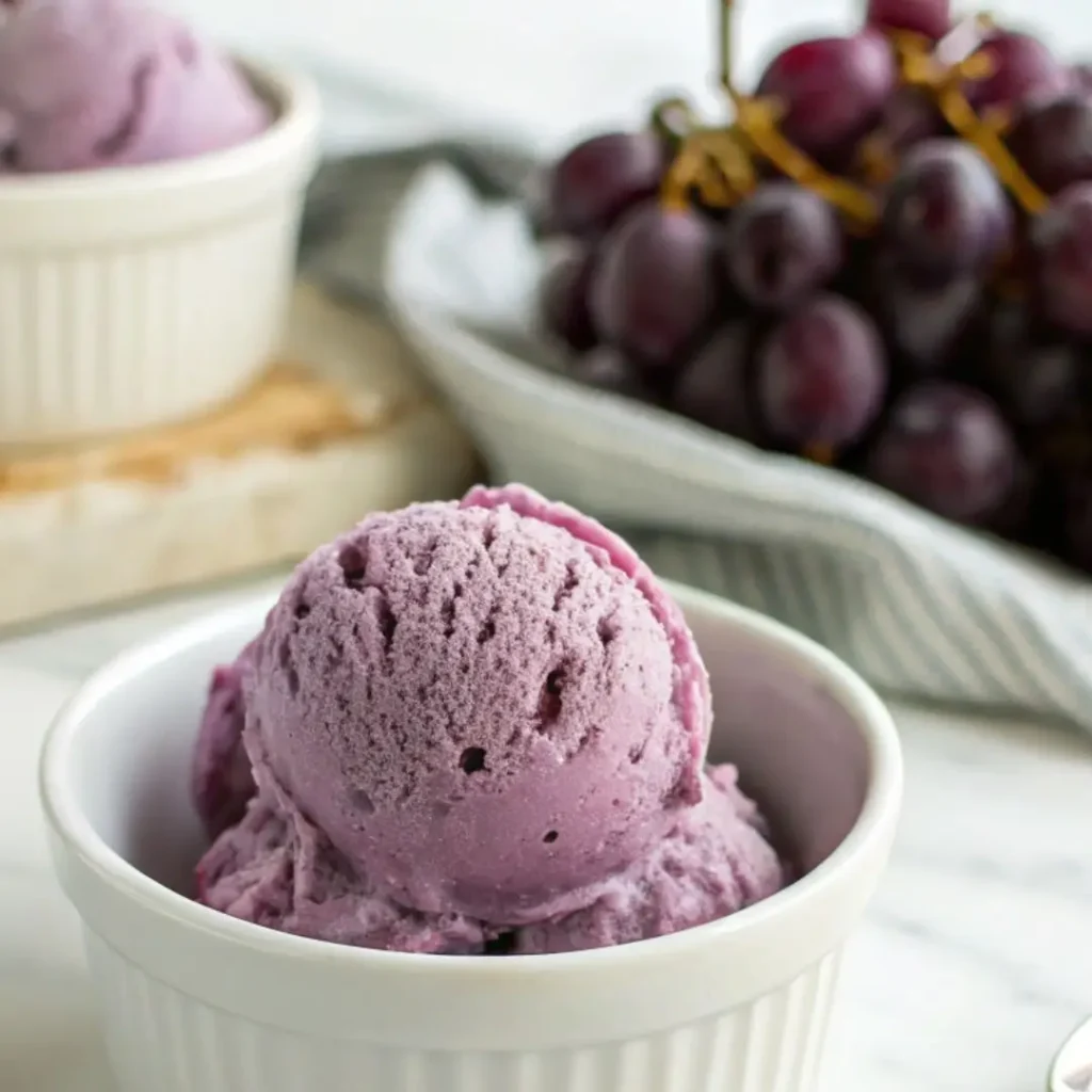 A nostalgic scoop of homemade grape ice cream recipe in a white bowl with Concord grapes, highlighting its creamy texture and vintage appeal.