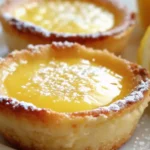 Close-up of baby lemon impossible pies with golden edges and a creamy lemon filling, topped with powdered sugar and sliced lemon in the background.