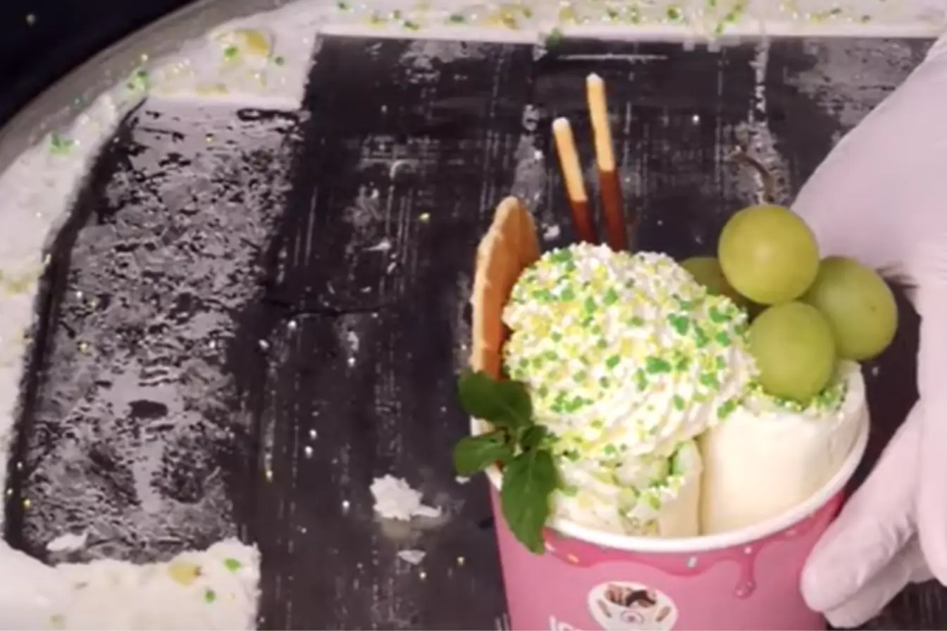 Grape ice cream rolls topped with whipped cream, green grapes, and sprinkles in a pink cup on a cold ice plate.
