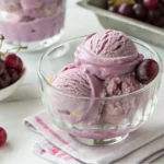 Three scoops of viral grape ice cream in a glass bowl, surrounded by fresh Concord grapes on a white background.