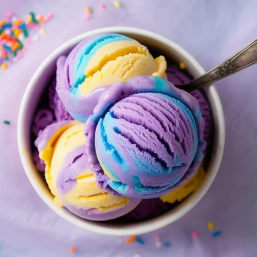 A close-up of a white bowl filled with scoops of Moon Mist Ice Cream in vibrant swirls of purple, blue, and yellow, with a vintage silver spoon and colorful sprinkles scattered on a pastel purple background.