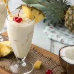 Creamy virgin piña colada in a tall glass with pineapple wedge, maraschino cherry, and striped straw, served with fresh pineapple slices and coconut.