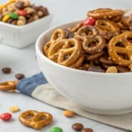 Butter toffee pretzels snack mix in a white bowl, with golden toffee-coated mini pretzels, nuts, and candies on a light background.