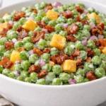 Classic pea salad with cheddar cubes, crispy bacon, and red onion in a white bowl.