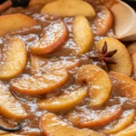 Close-up of stovetop apple pie filling with fresh apple slices, cinnamon sticks, and thick syrup in a cast iron skillet.
