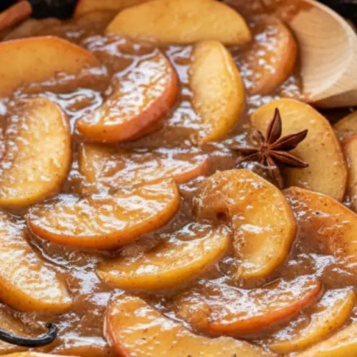 Close-up of stovetop apple pie filling with fresh apple slices, cinnamon sticks, and thick syrup in a cast iron skillet.