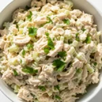Healthy chicken salad recipe in a white bowl with shredded chicken, diced celery, parsley, and a creamy Greek yogurt dressing.