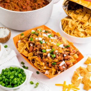 Frito pie recipe served in an orange tray with corn chips, beef chili, melted cheddar cheese, diced onion, and green onions, surrounded by chili, Fritos, and toppings.