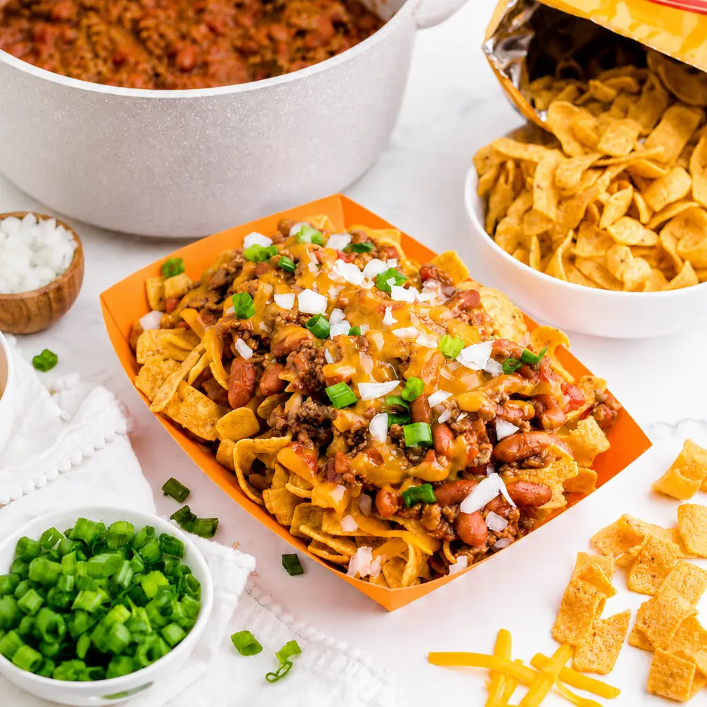 Frito pie recipe served in an orange tray with corn chips, beef chili, melted cheddar cheese, diced onion, and green onions, surrounded by chili, Fritos, and toppings.