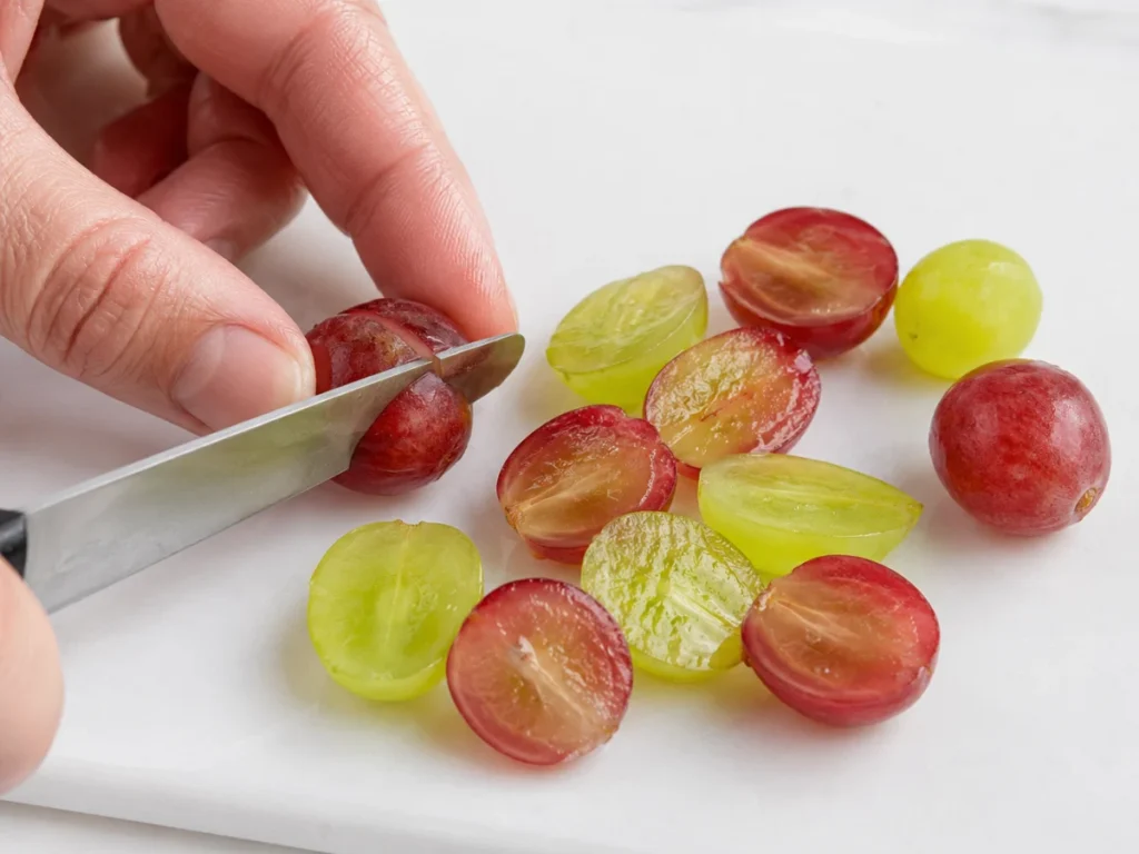 Red and green seedless grapes being halved with a small knife on a white cutting board