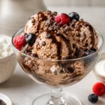 High protein ice cream scooped into a glass bowl with chocolate drizzle, berries, and crunchy mix-ins.